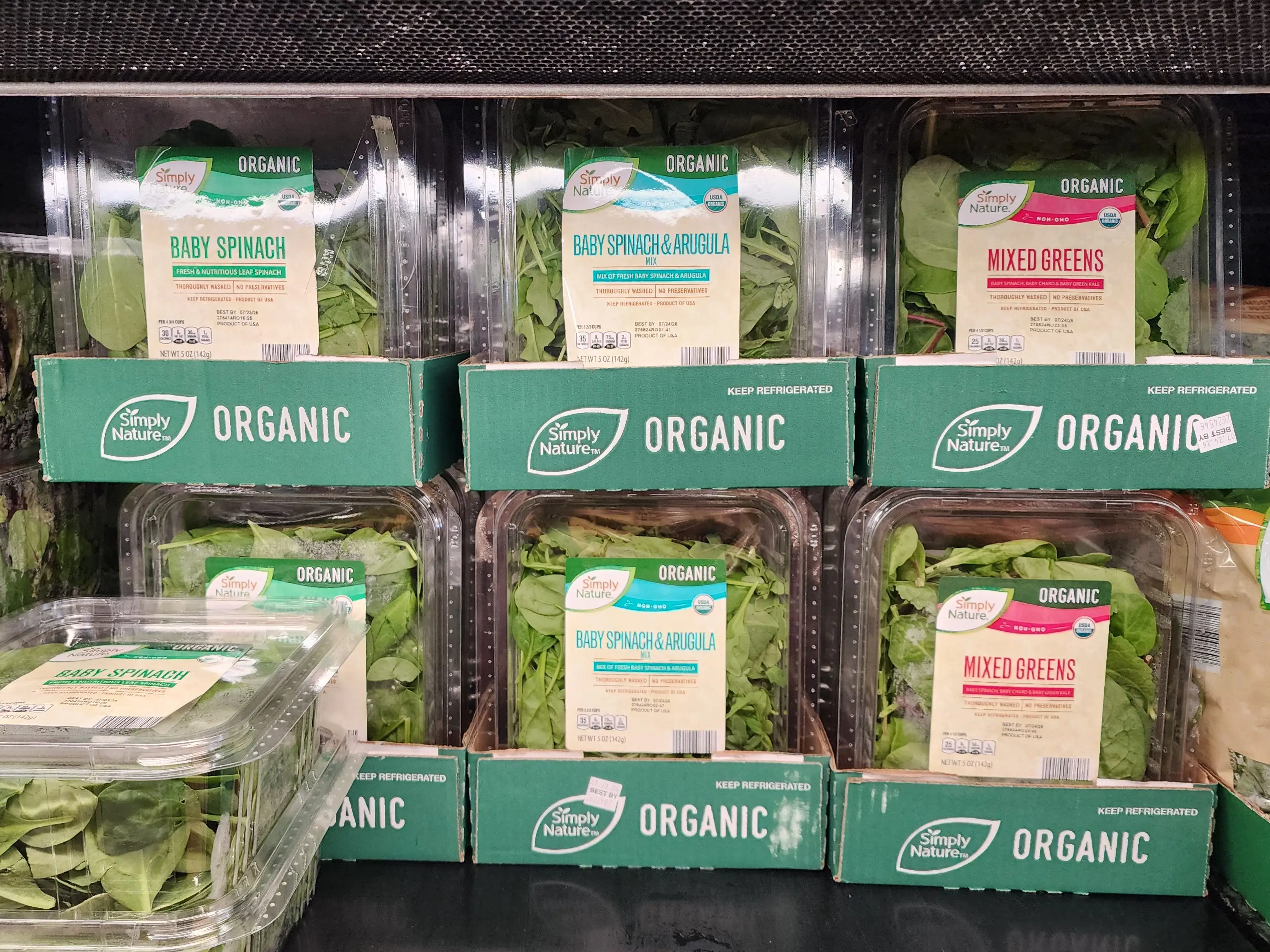 Several boxes of leafy greens, including baby spinach, arugula, and mixed greens, sitting on a shelf at Aldi.
