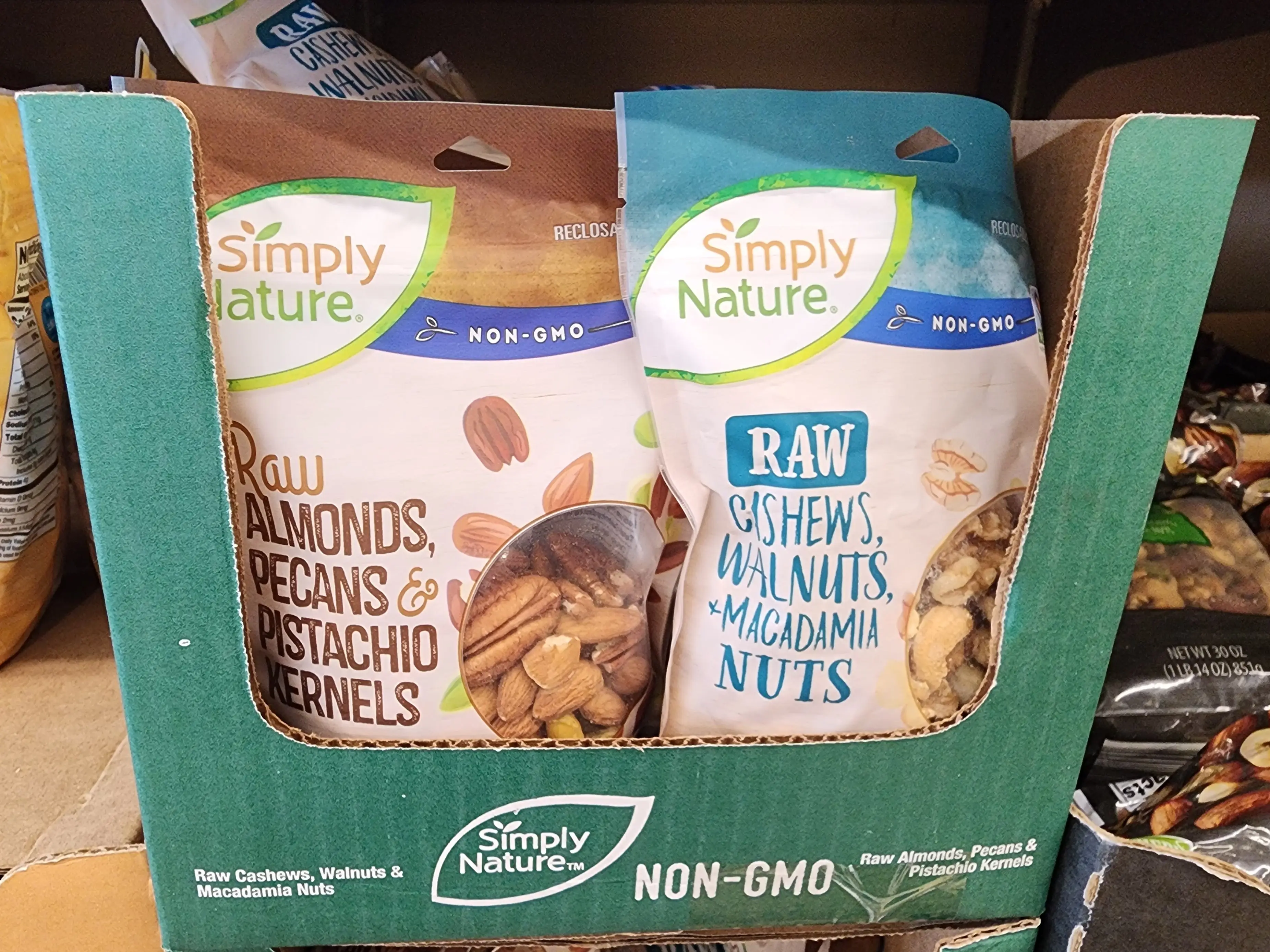 A bag of Simply Nature raw almonds, pecans, and pistachio kernels, and a bag of Simply Nature raw cashews, walnuts, and macadamia nuts sit in a cardboard box at Aldi.