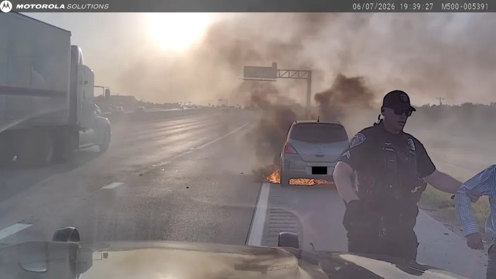 A police officer assisting a person near a burning car on the side of a highway.