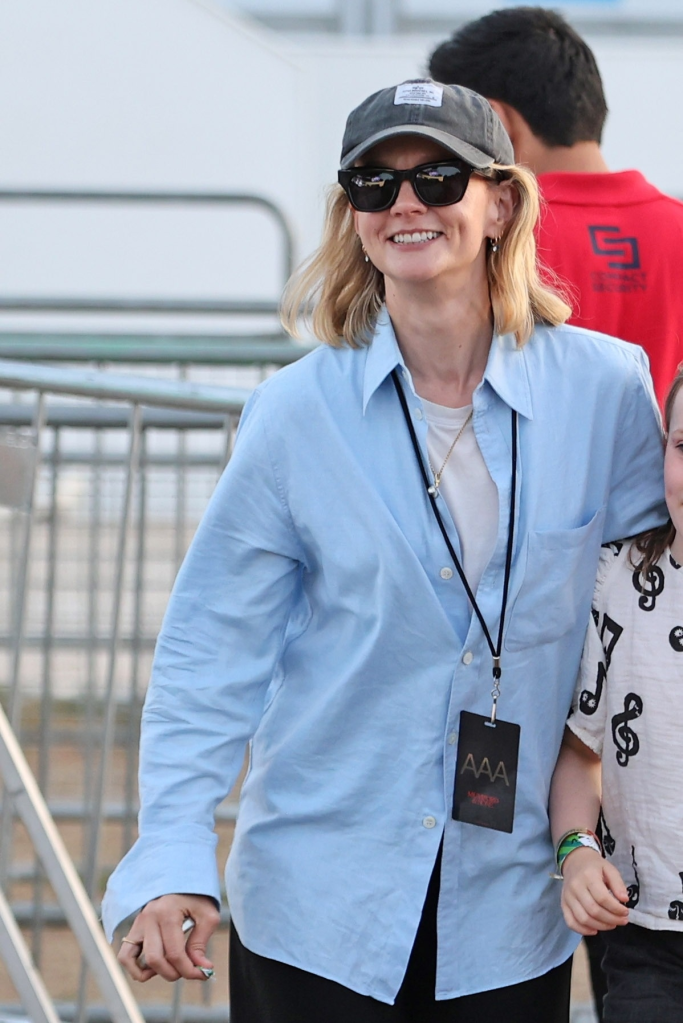 Carey Mulligan smiles while wearing a light blue button-down shirt, a white t-shirt, black sunglasses, and a gray baseball cap with an 
