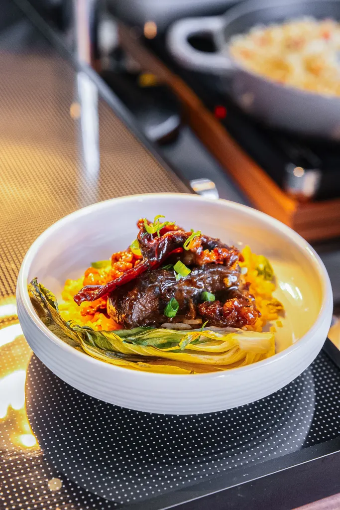 White bowl of meat with bok choy and peppers on a black and white polka-dot surface.