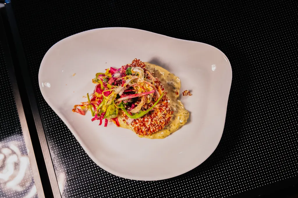 A white plate with a dish featuring a colorful salad over a creamy sauce, garnished with red quinoa.