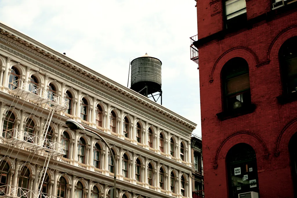 A large, cream-colored building with ornate architectural details and a fire escape, with a water tower on the roof and a red brick building to the right.