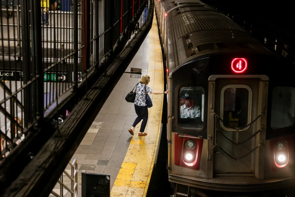 A woman in a floral shirt and dark pants stands on a subway platform next to a train with a red