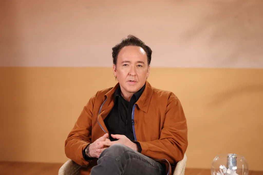 Actor John Cusack in a brown jacket, black shirt, and gray jeans, seated with hands clasped.