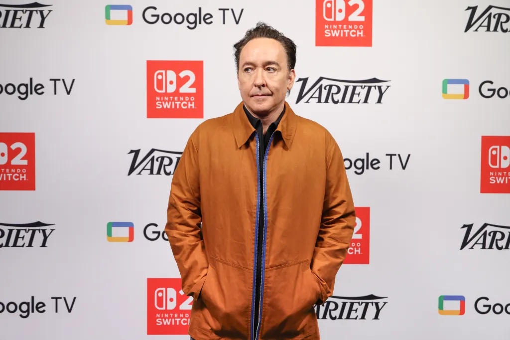 John Cusack posing for a photo in front of a white backdrop with 