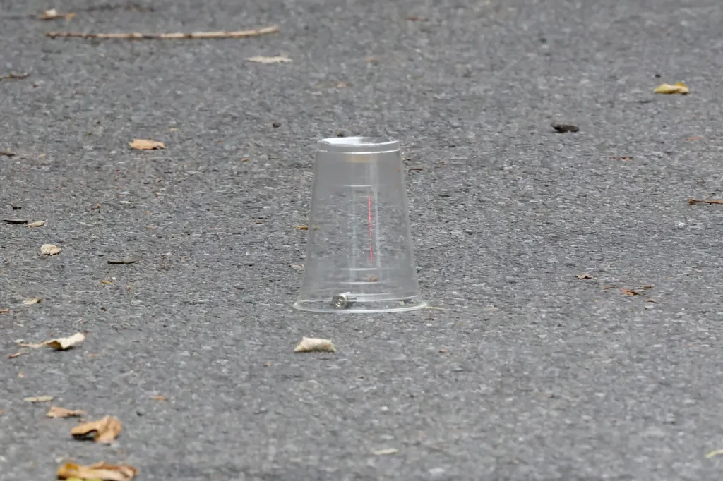 A clear plastic cup on asphalt covers a bullet casing, typical of a crime scene.