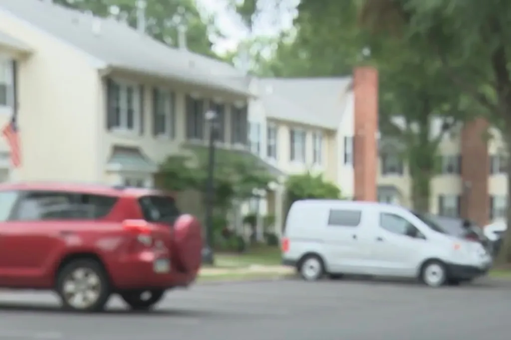 A blurred image of a street with several houses in the background and a red SUV and a white van in the foreground.