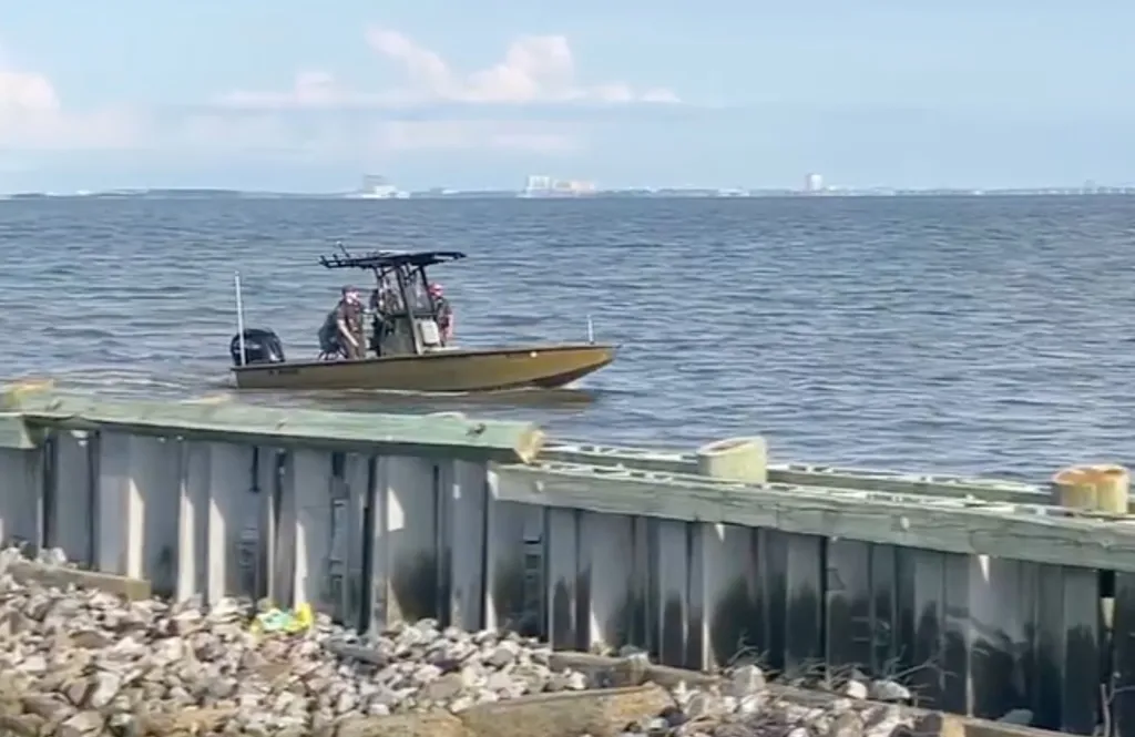 A brown boat with two people on board floating on the ocean, with a city skyline in the background.