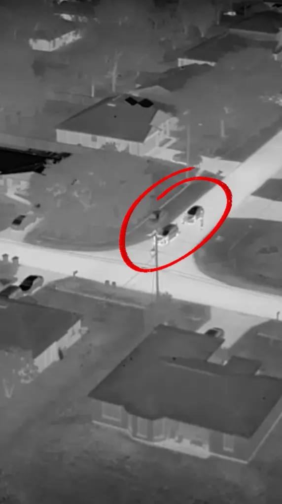 Aerial thermal image of a suburban street with houses and trees, showing two cars highlighted in a red oval.