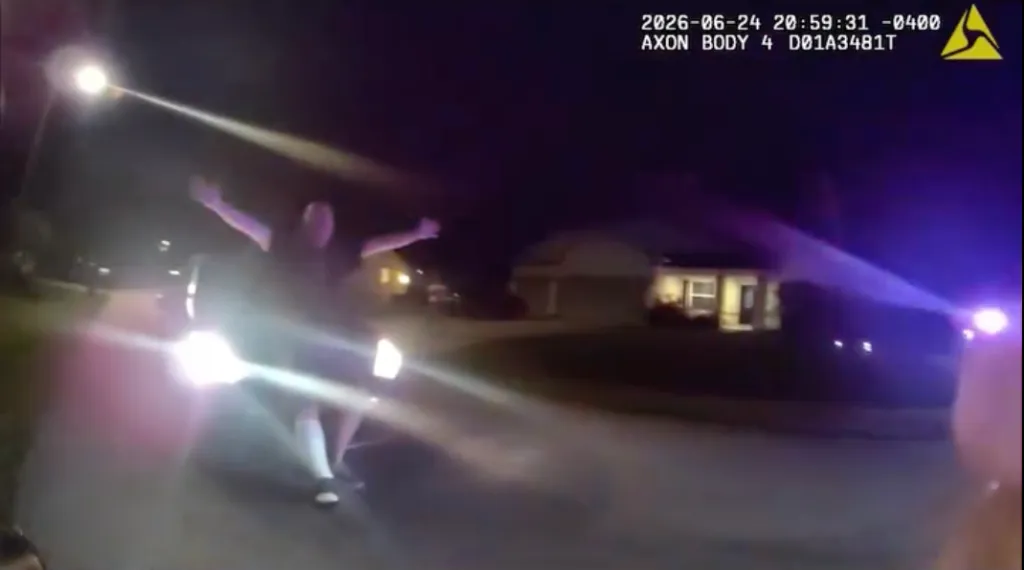 A person stands in the street with their arms outstretched, facing a vehicle with its headlights and flashing blue lights on, at night.