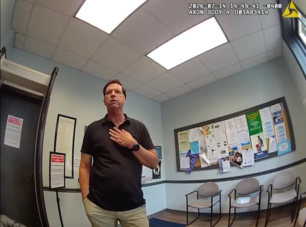 A man in a black polo shirt and khakis standing in a waiting room with his hand on his chest.