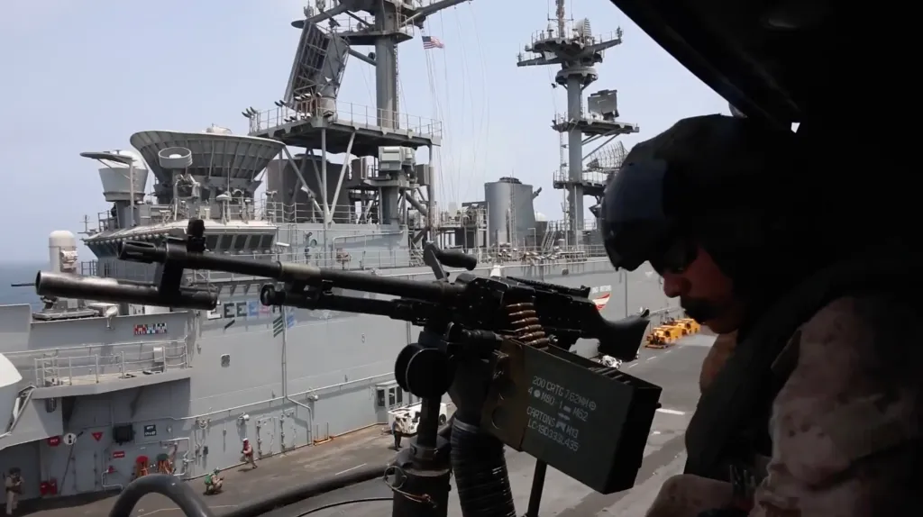 A machine gun mounted on a helicopter with a soldier next to it, overlooking a ship.