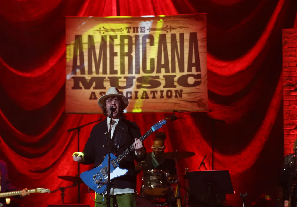 Aaron Lee Tasjan performs on stage, wearing a fedora and glasses, playing a blue electric guitar in front of a red curtain and 