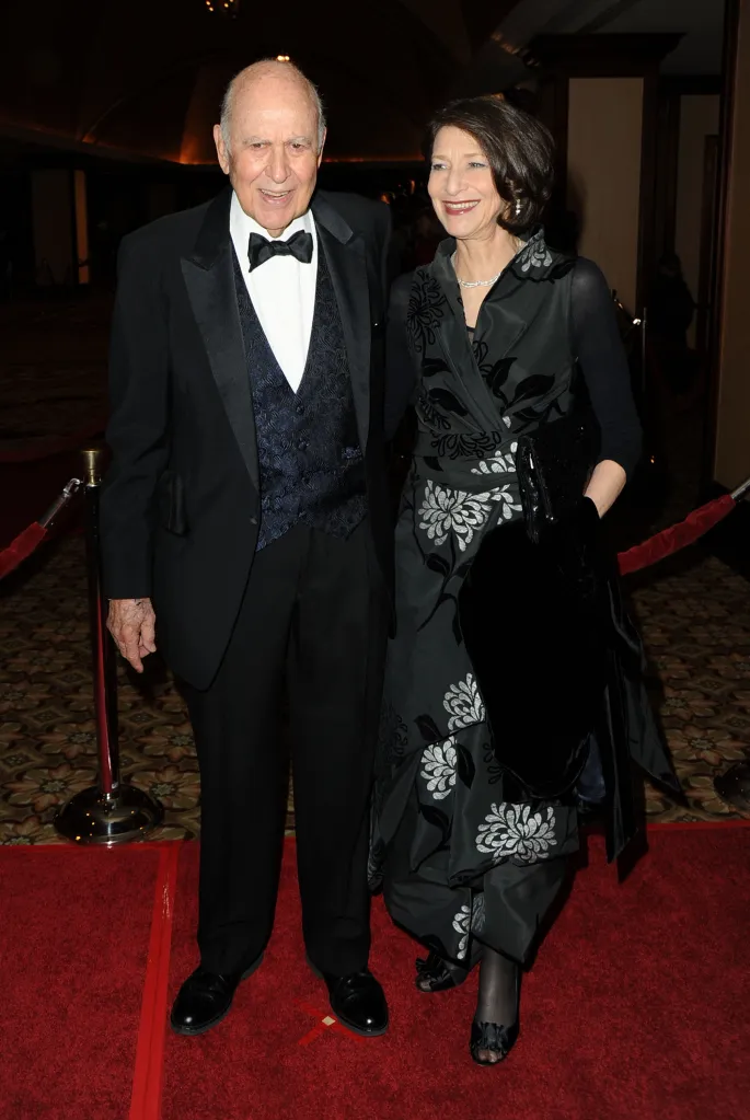 Carl Reiner in a tuxedo and his daughter Annie Reiner in a floral dress on a red carpet.