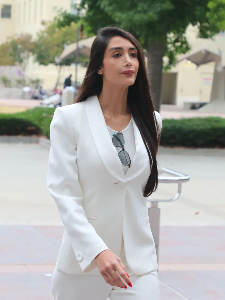 Adva Lavie, a woman with long dark hair, wearing a white pantsuit and sunglasses tucked into her top, walks forward.