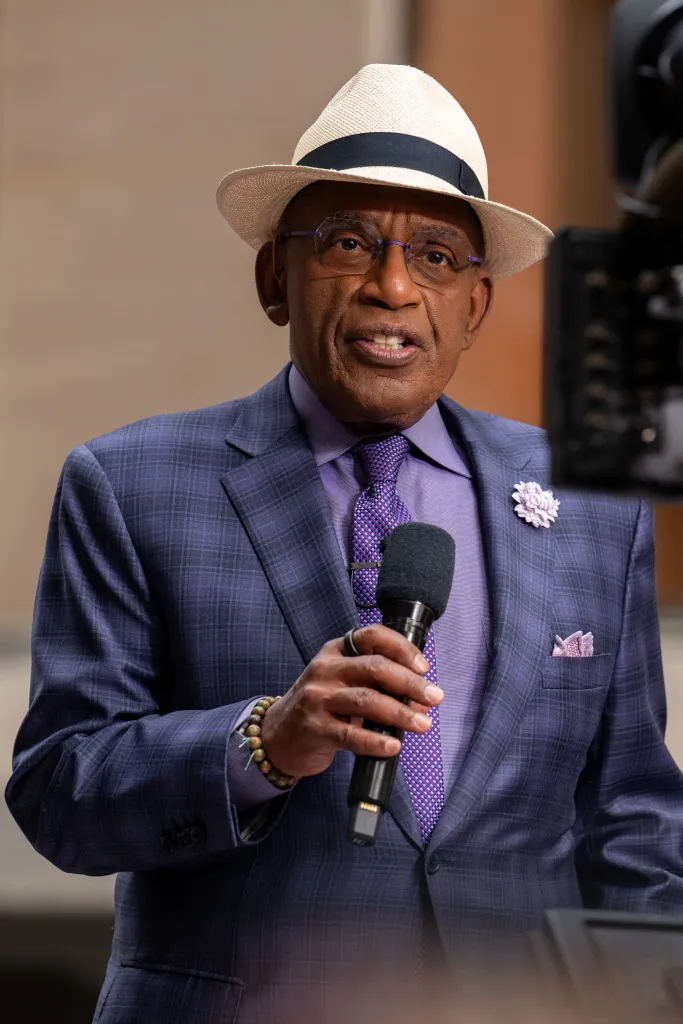 Al Roker, wearing a hat, glasses, and a purple plaid suit, speaks into a microphone.