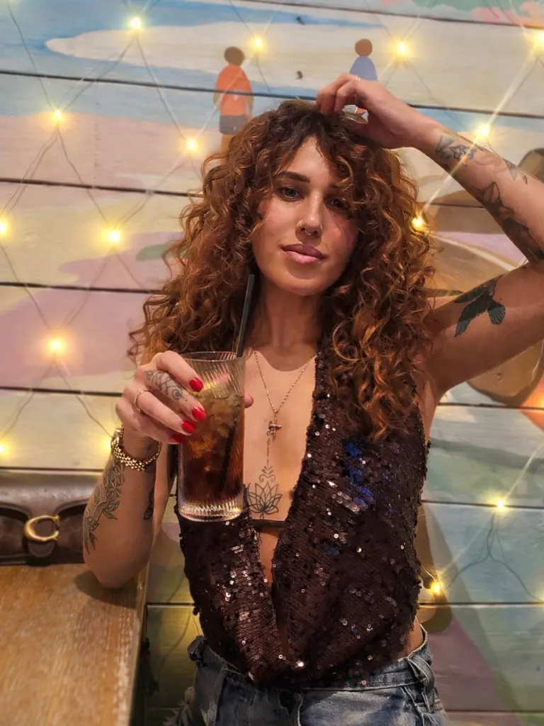 A woman with curly brown hair, wearing a sequined top and jeans, holding a drink and touching her hair, in front of a colorful wall with string lights.