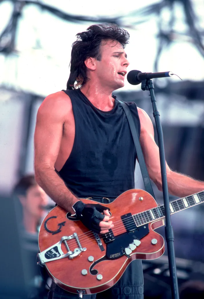 Rick Springfield performing onstage at Live Aid, playing a guitar, with sweat visible on his face and chest.