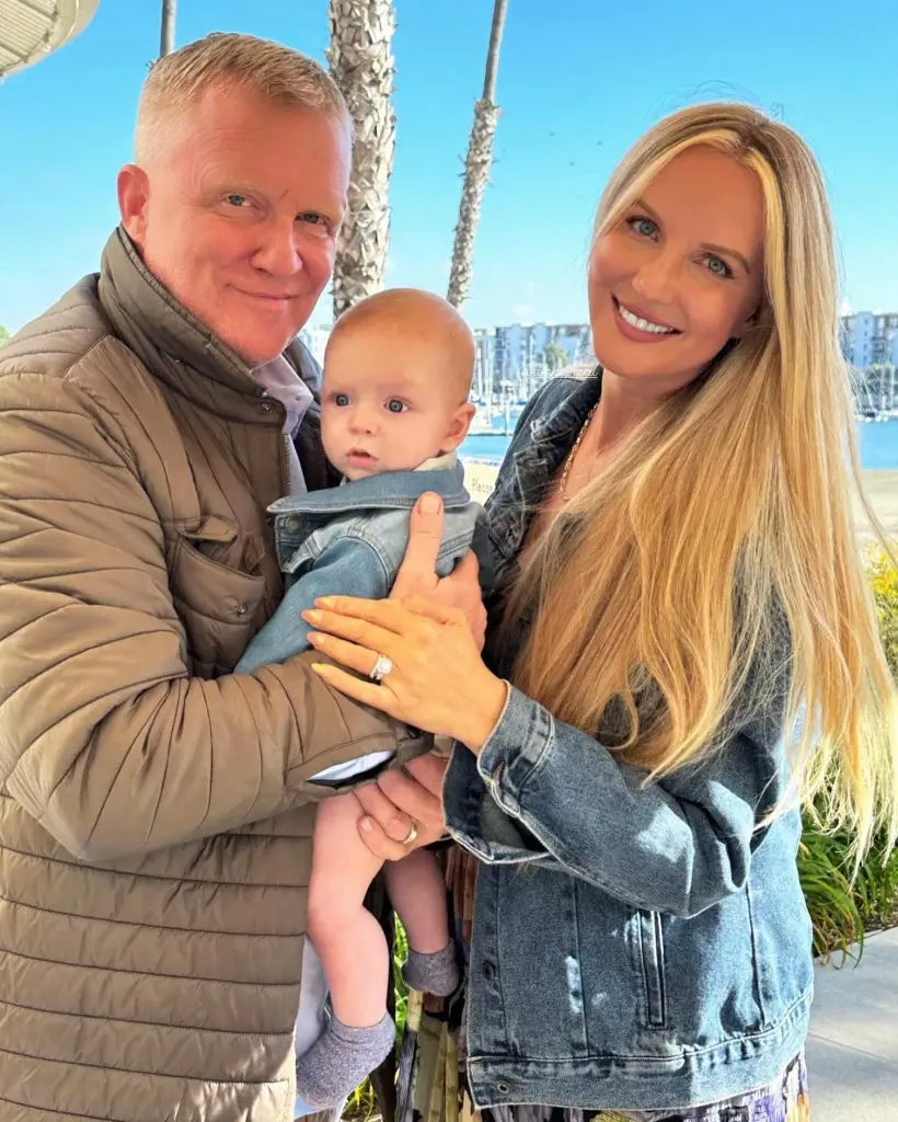 Anthony Michael Hall, his wife, and their baby smile for a photo with a marina in the background.