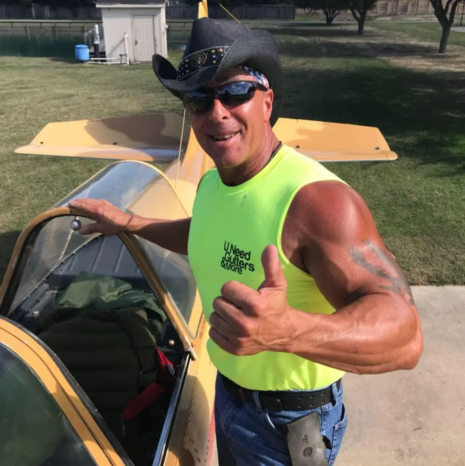 Anthony Soraci in a black cowboy hat and sunglasses, giving a thumbs up next to a small yellow plane.