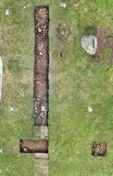 An aerial view of an archaeological excavation site with a long trench revealing historic remnants, and two smaller excavation squares in grassy ground.