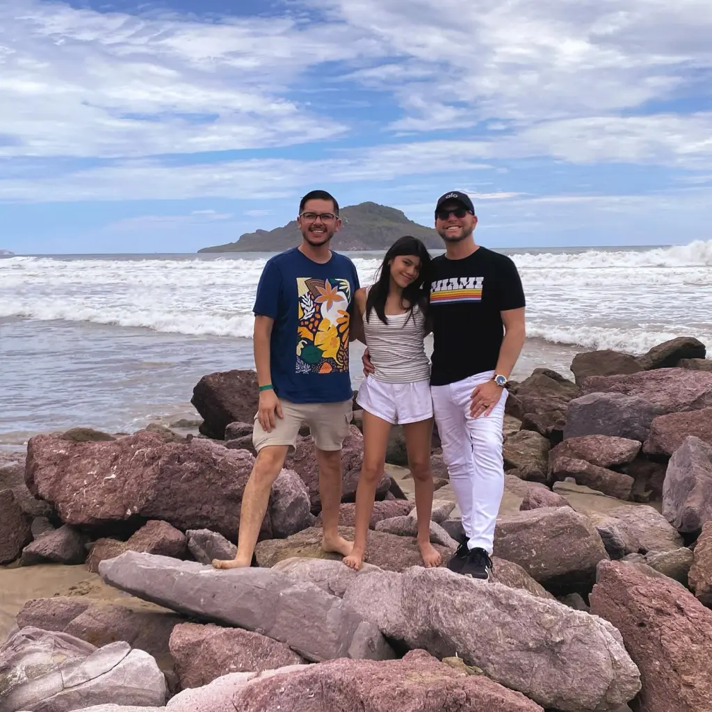 Armando Niedermeier-Rubio posing with his daughter and another man on rocks by the ocean.