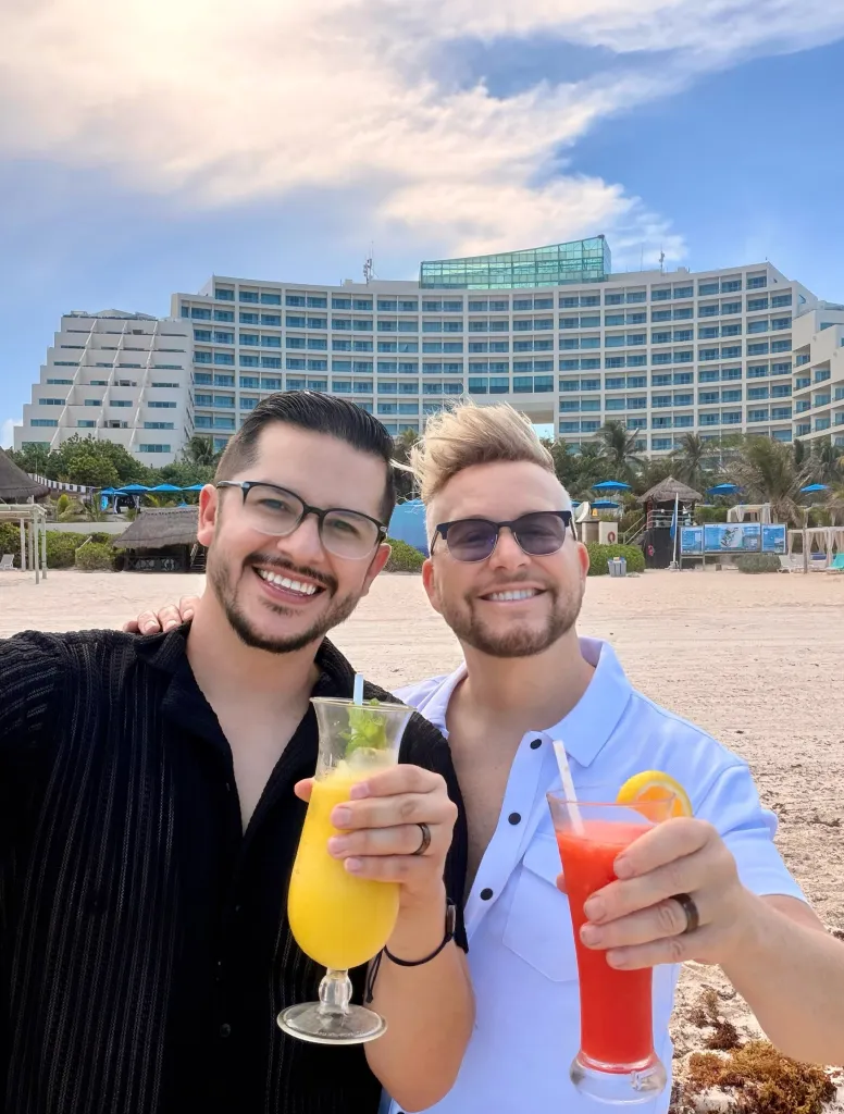 Armando Rubio and Kenneth Niedermeier smiling and holding tropical drinks on a beach.