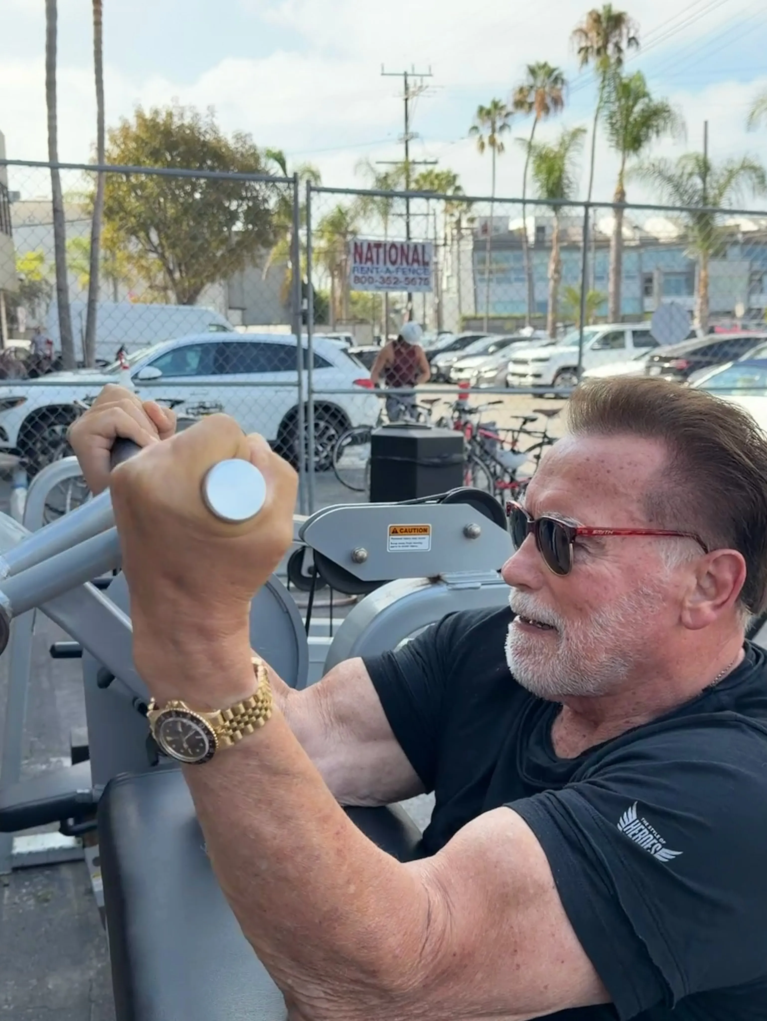 Arnold Schwarzenegger exercising outdoors, wearing sunglasses and a gold watch.