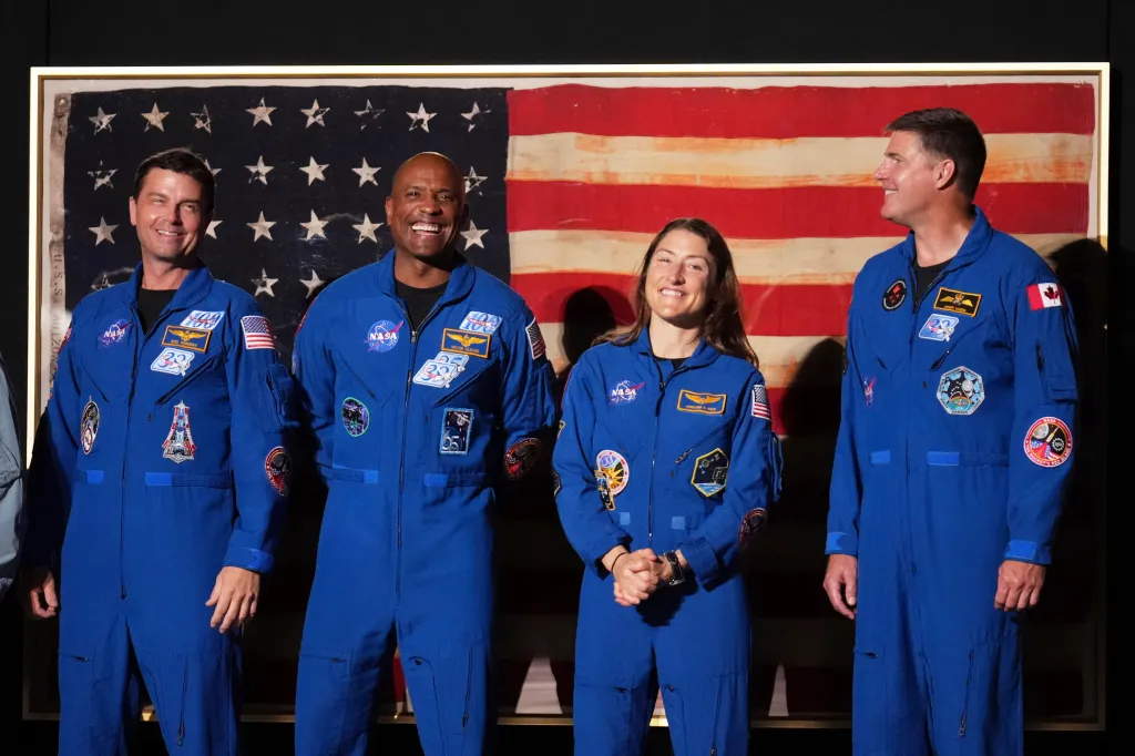 Artemis II crew members Reid Wiseman, Victor Glover, Christina Koch, and Jeremy Hansen standing in front of a US flag.