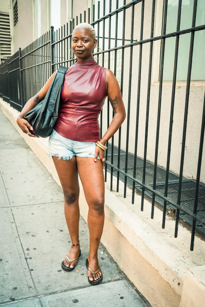 Ashley Renée standing on W Houston St, Manhattan, NY, wearing a maroon top, distressed shorts, and flip-flops.
