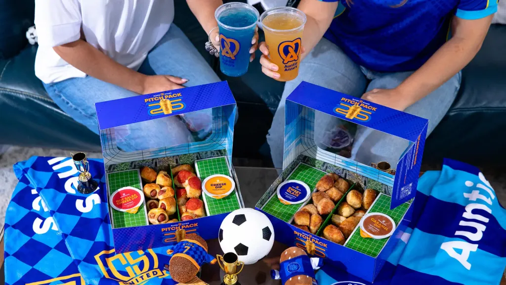 Two people holding Auntie Anne's pretzel drinks and two Pitch Pack boxes filled with pretzel bites, surrounded by soccer-themed merchandise.