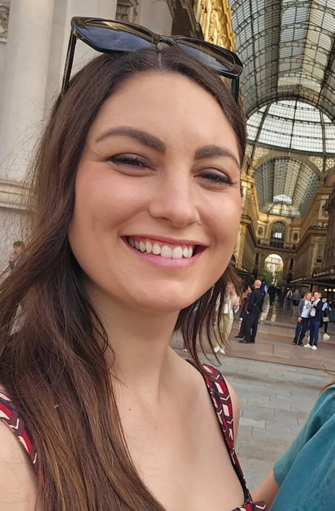 A woman with long brown hair and sunglasses on her head smiles while standing in front of a grand arched building with a glass ceiling.