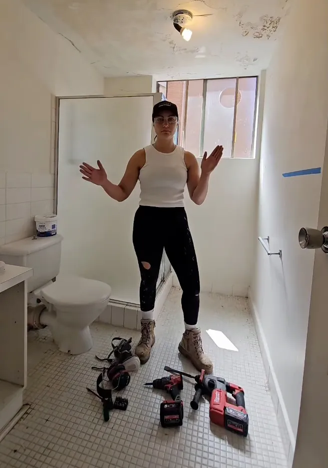 Woman in work clothes, surrounded by tools, showing the state of a dilapidated room.