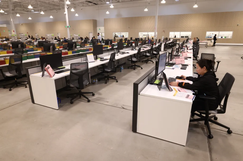 A worker processes ballots at a computer in a large room filled with similar workstations.