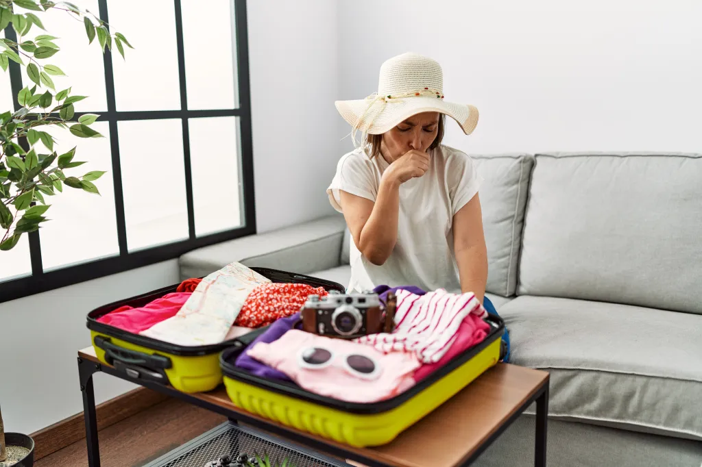 A middle-aged Hispanic woman, wearing a sunhat and seated on a sofa, coughs into her hand while packing a suitcase with summer clothes.
