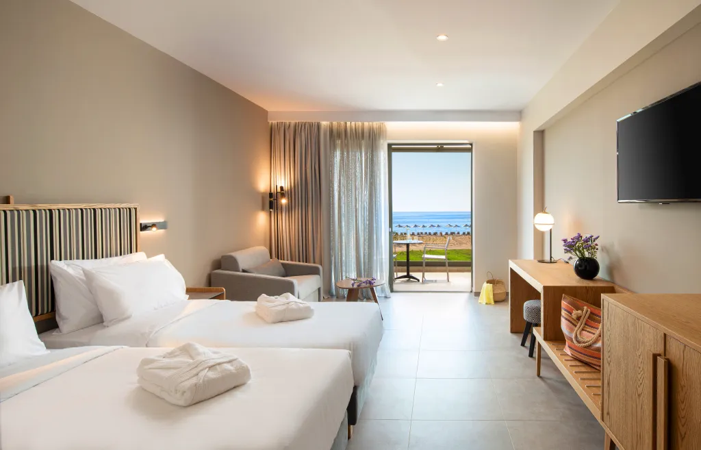 Modern double hotel room with two beds, a seating area, a desk, and a balcony with a sea view.