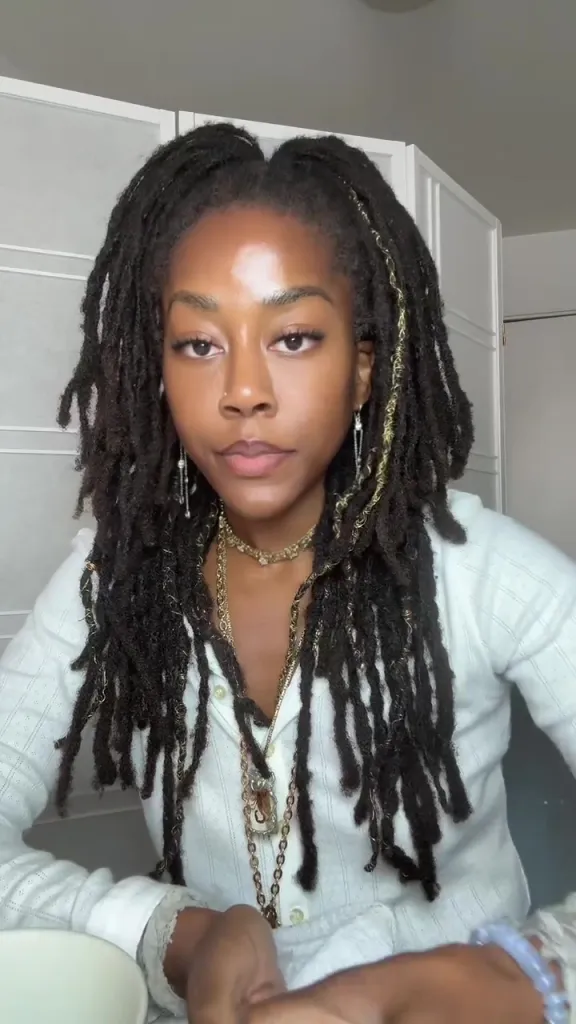 A woman with long dreadlocks and gold jewelry wearing a white shirt.