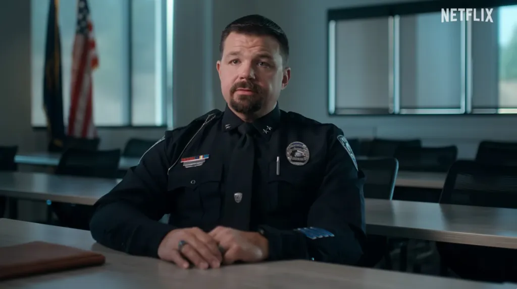 A police officer sitting at a table with an American flag in the background.