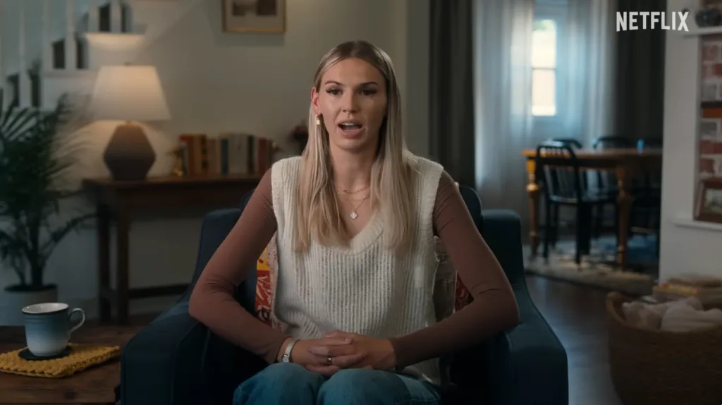 A woman in a cream sweater vest over a brown long-sleeved shirt sitting in a dark blue armchair in a living room, speaking.