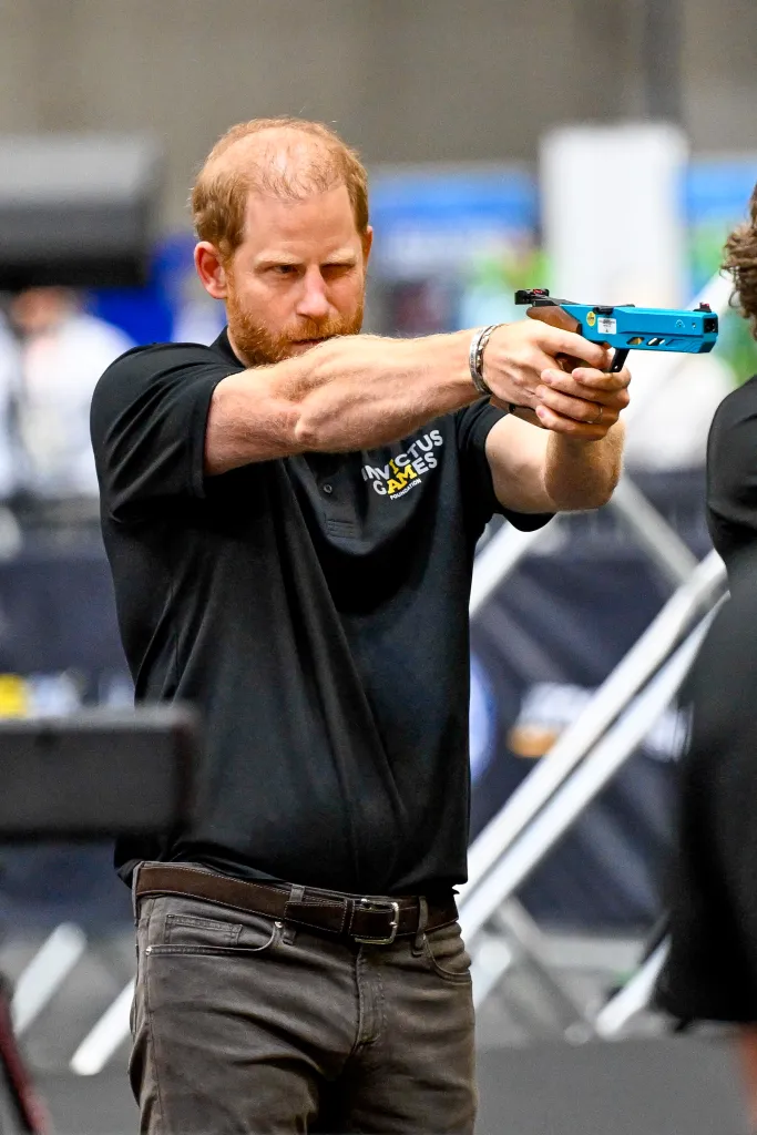 Prince Harry aiming a blue air pistol, wearing a black polo shirt with 