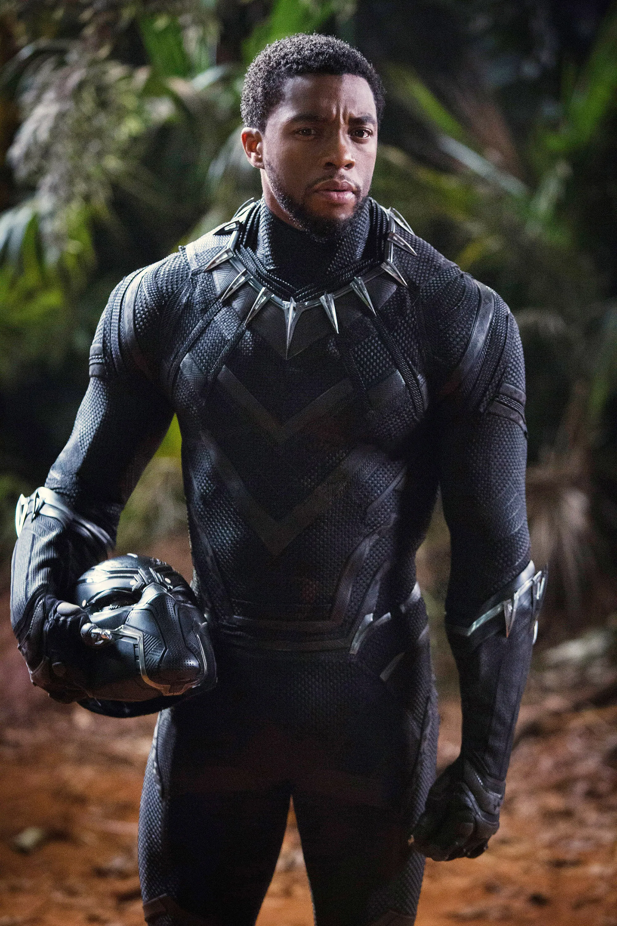 Chadwick Boseman as Black Panther, holding his mask, in a black textured suit with a spiked necklace.