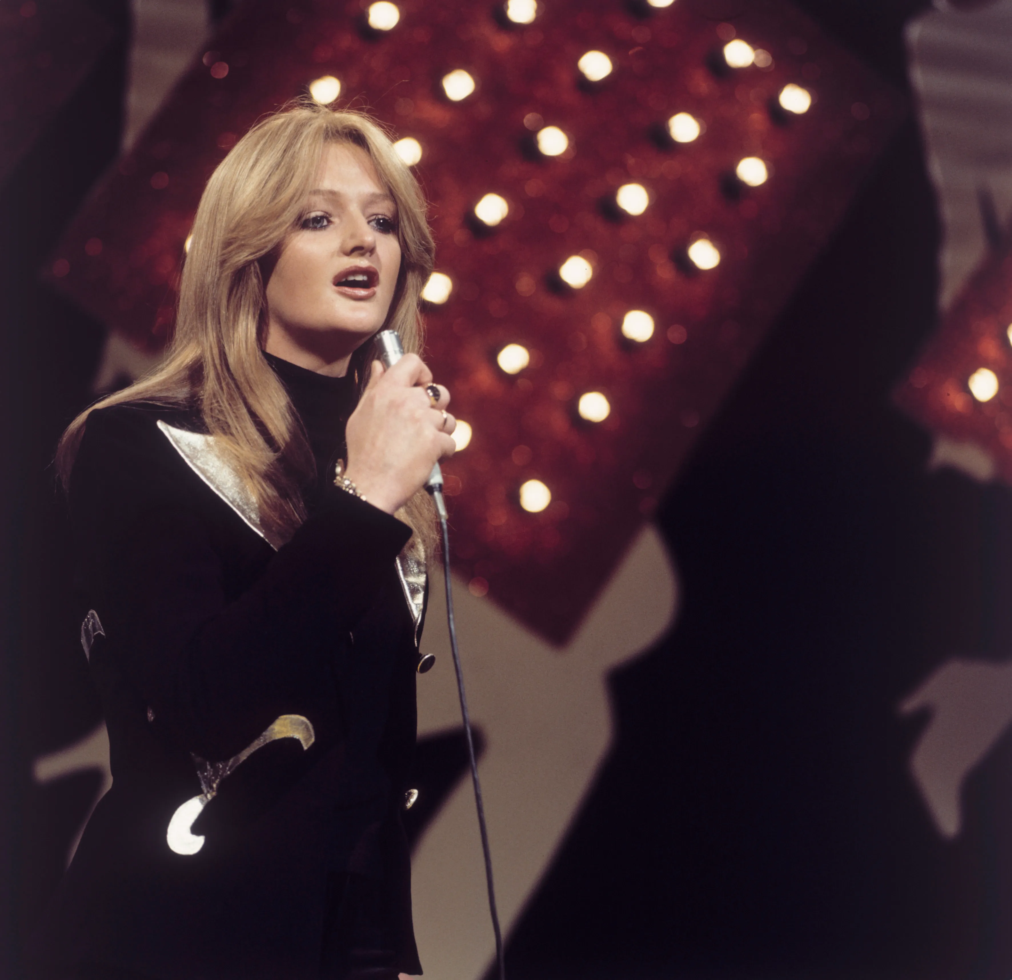 Bonnie Tyler singing into a microphone on stage, with a red backdrop featuring bright, circular lights.