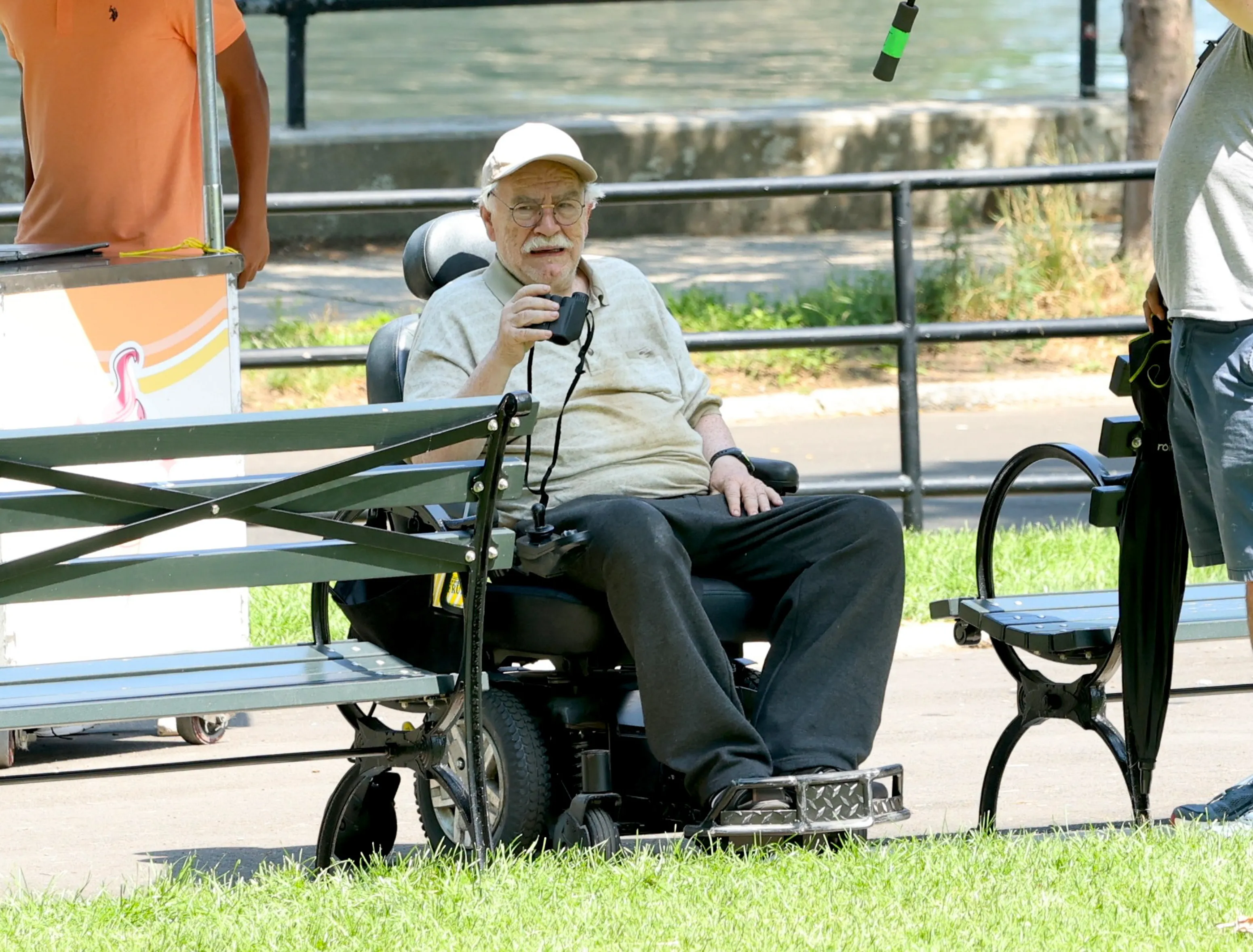Brian Cox in a white baseball cap and light shirt, seated in a motorized wheelchair and holding binoculars.