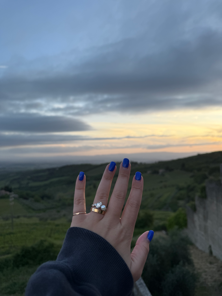 A hand with bright blue nail polish and rings against a sunset landscape.