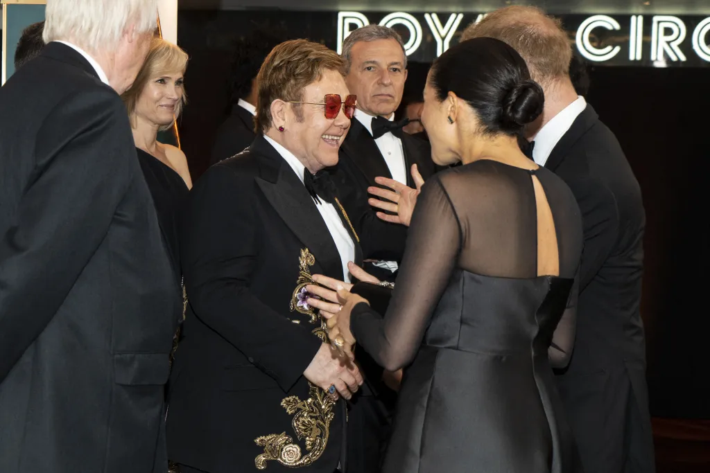Elton John, in a black suit with gold brocade, and a black bow tie and sunglasses, chatting with Meghan, Duchess of Sussex, in a black gown, and Prince Harry, Duke of Sussex, in a black tuxedo, at the European premiere of The Lion King.