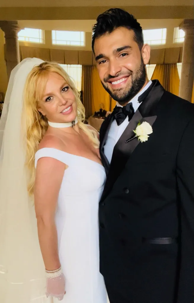Britney Spears, in a white wedding gown and white veil, and Sam Asghari, in a black tuxedo, smiling at their wedding.