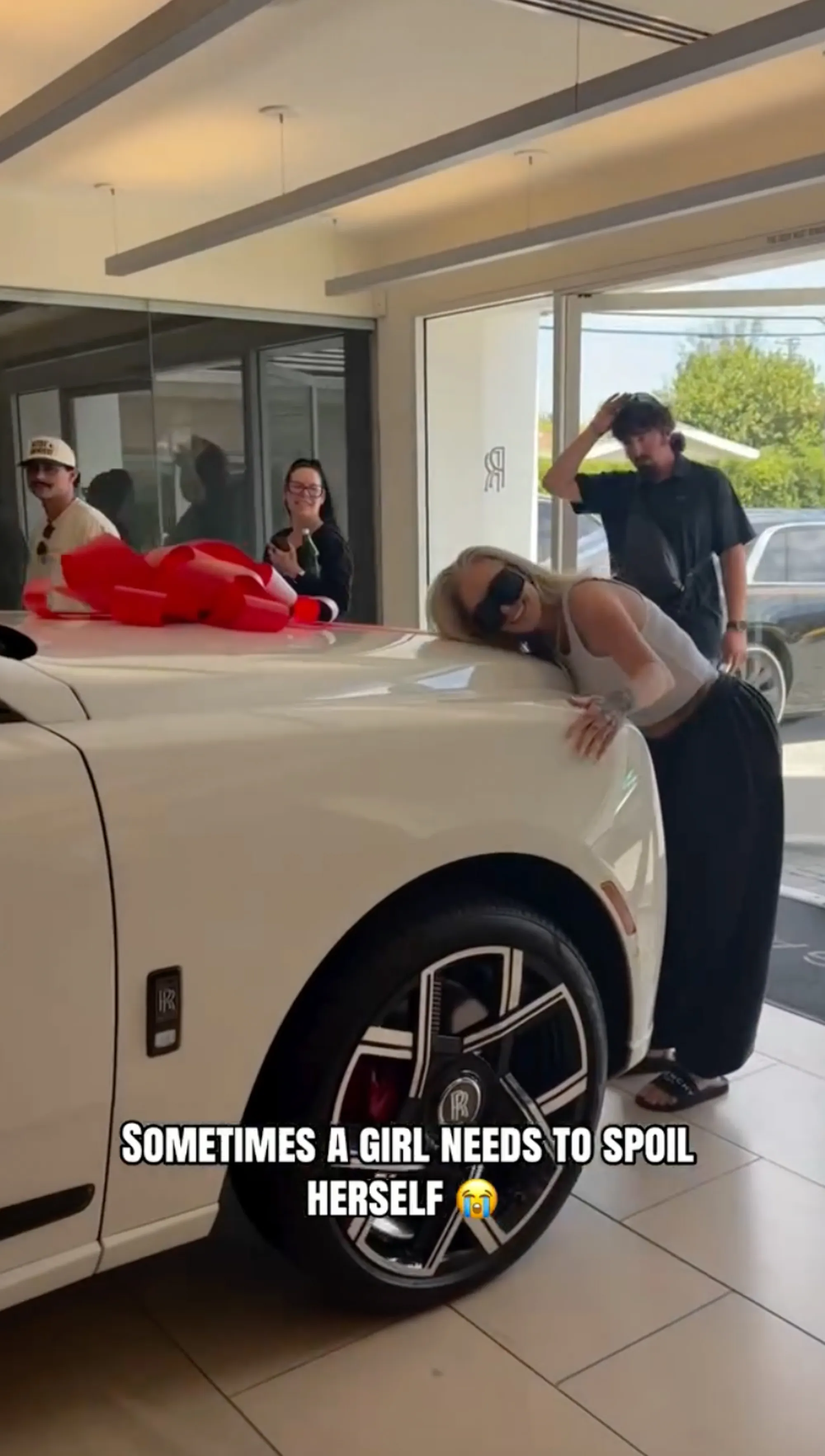 Bunnie Xo leaning on a white Rolls-Royce with a red bow, surrounded by people in a dealership.