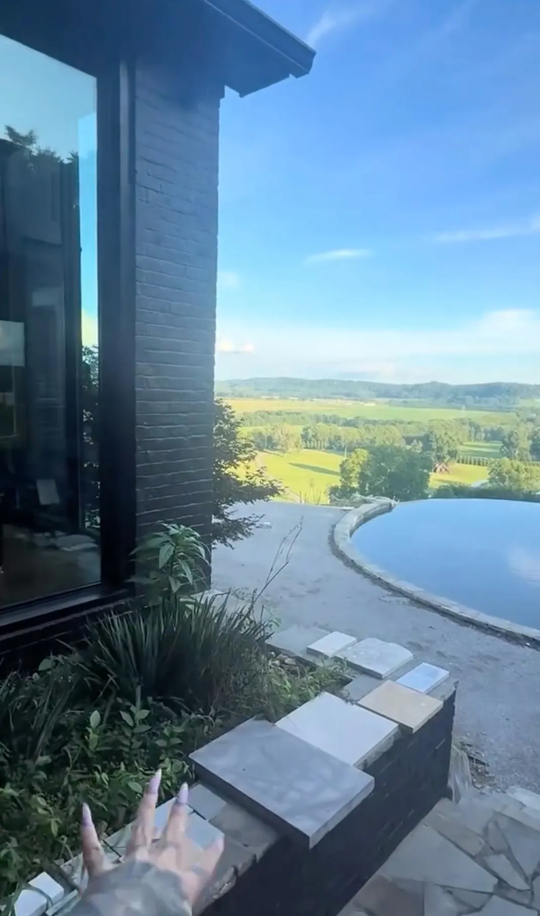 A house with large windows overlooks a wide green landscape, and a curved infinity pool is visible on the right.