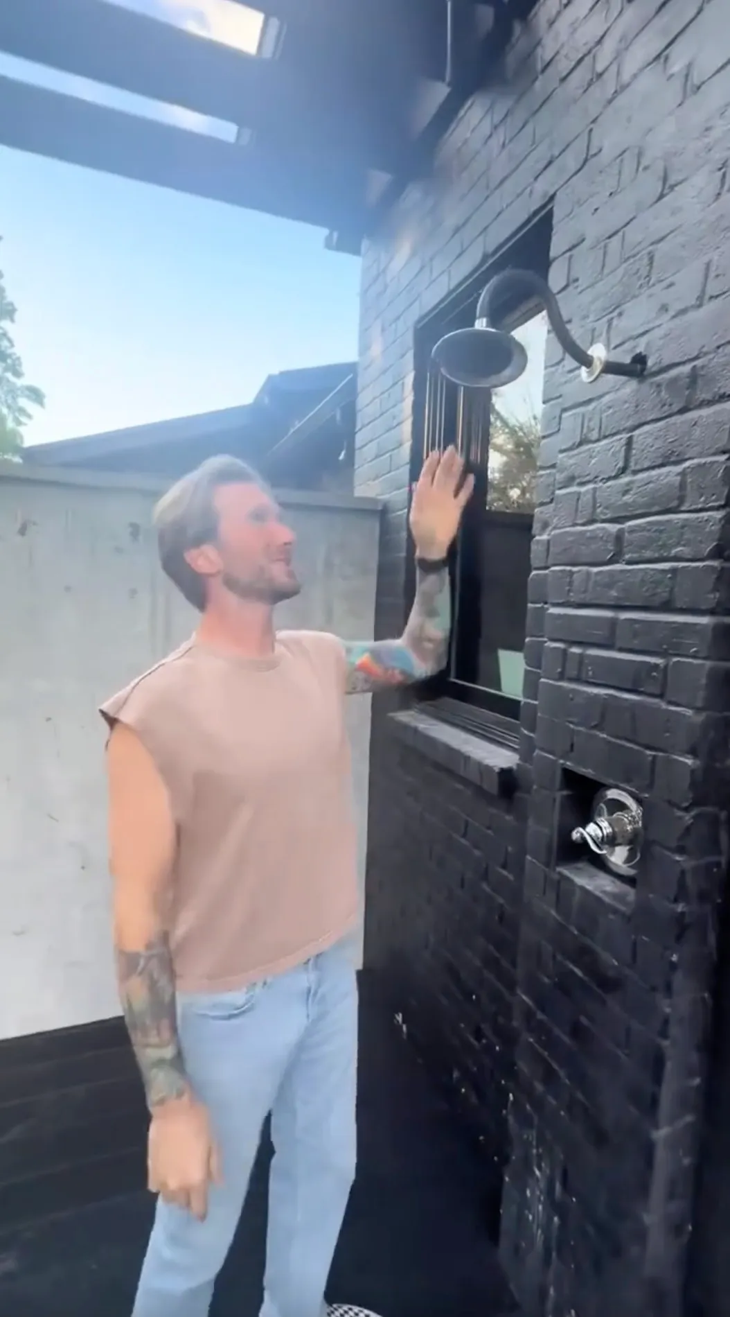Man in a tan shirt and light-wash jeans gesturing to an outdoor shower on a black brick wall.